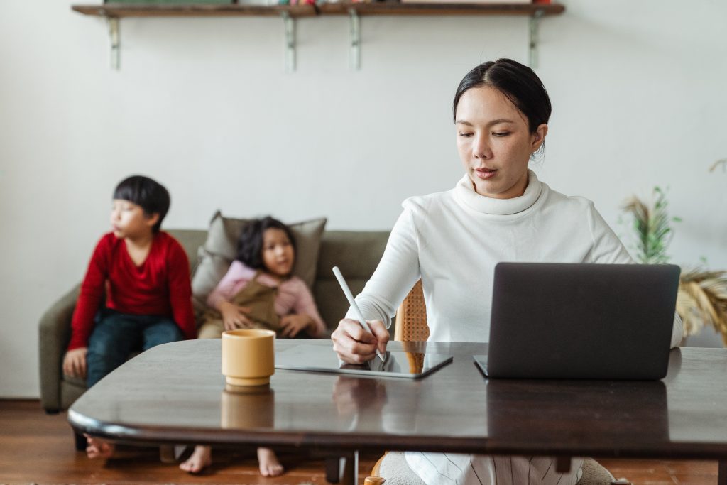 A woman sits at her computer taking notes while her two children are on the couch behind her